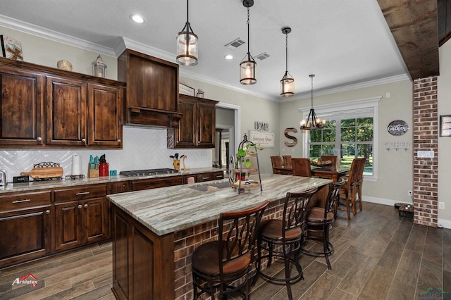 kitchen with decorative light fixtures, light stone counters, stainless steel gas stovetop, and an island with sink