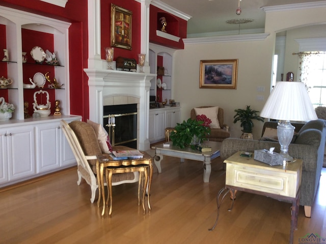 living room featuring a tiled fireplace, built in features, ornamental molding, and light hardwood / wood-style flooring