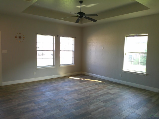 empty room with a raised ceiling, plenty of natural light, dark wood-type flooring, and ceiling fan