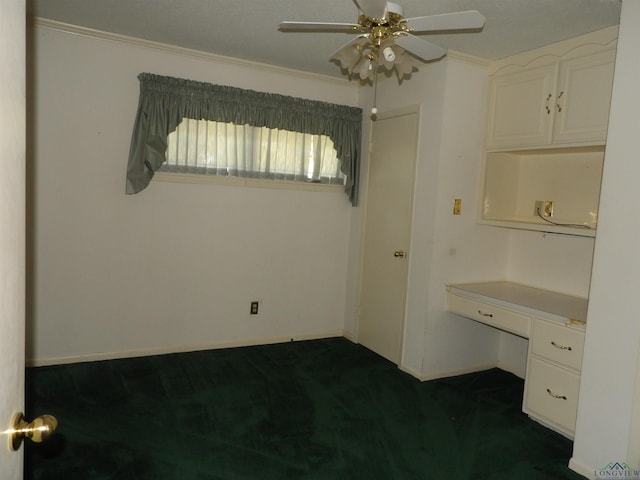 spare room with dark colored carpet, built in desk, ceiling fan, and ornamental molding