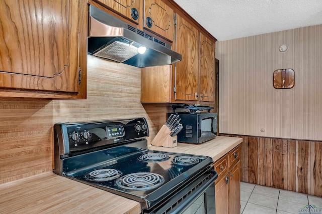 kitchen featuring black appliances, brown cabinets, under cabinet range hood, and wooden walls