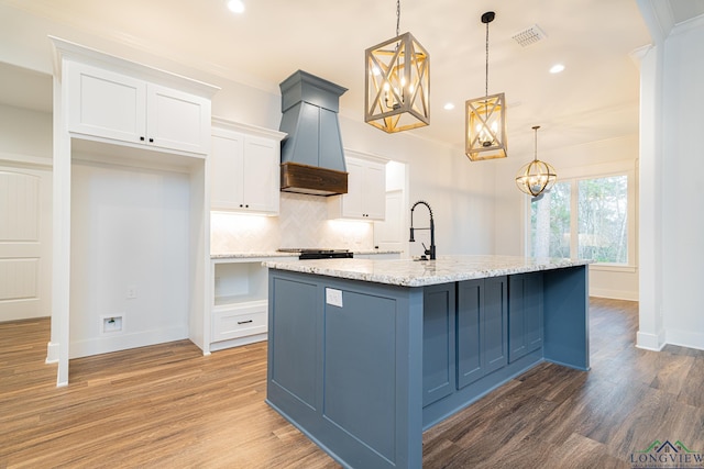 kitchen featuring light stone countertops, white cabinets, hanging light fixtures, a kitchen island with sink, and custom range hood