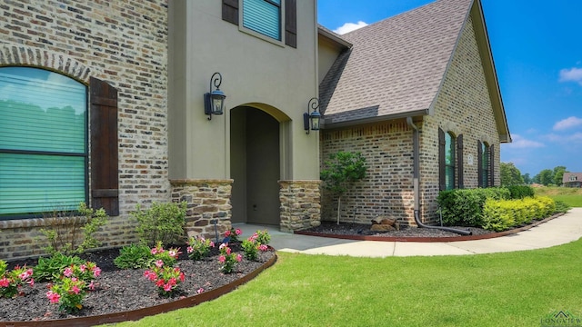 view of exterior entry featuring stucco siding, stone siding, a yard, roof with shingles, and brick siding