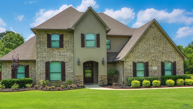 french country style house with a shingled roof, a front yard, and stucco siding