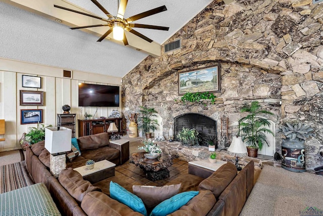 living room featuring a textured ceiling, a wood stove, ceiling fan, beamed ceiling, and a stone fireplace