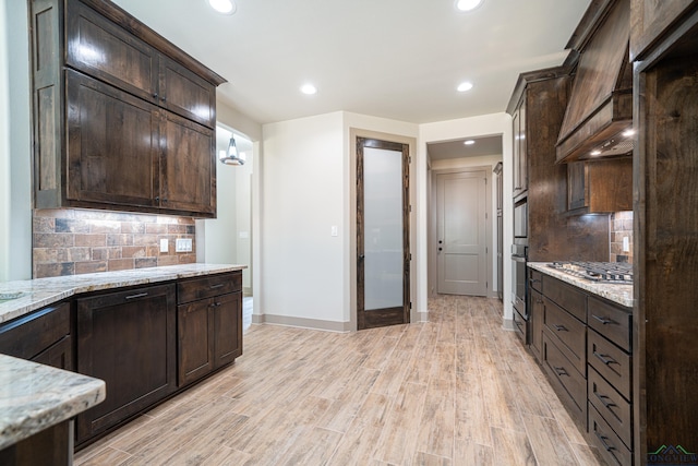 kitchen with oven, decorative backsplash, custom range hood, light stone counters, and dark brown cabinetry