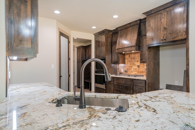 kitchen featuring light stone countertops, backsplash, premium range hood, dark brown cabinetry, and sink