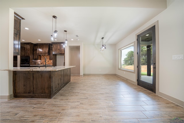 kitchen with custom exhaust hood, sink, hanging light fixtures, light stone counters, and dark brown cabinetry