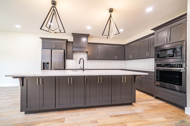 kitchen featuring light wood-type flooring, stainless steel appliances, a kitchen island with sink, sink, and hanging light fixtures