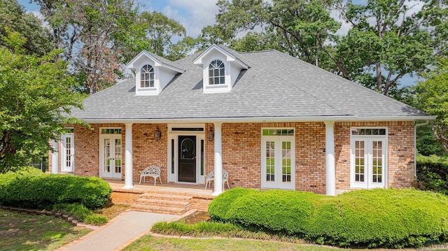 cape cod-style house featuring a porch, brick siding, french doors, and roof with shingles