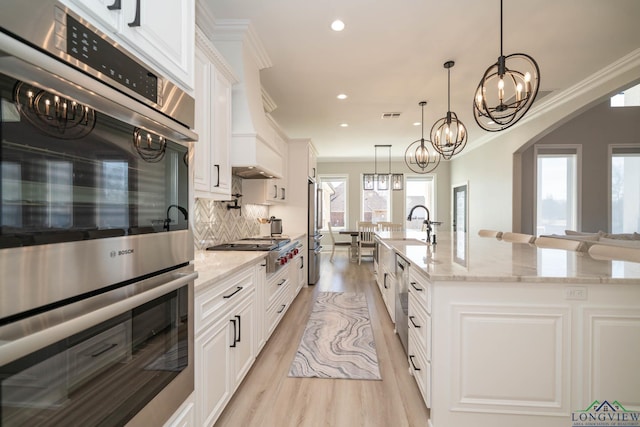 kitchen featuring pendant lighting, white cabinetry, stainless steel appliances, a kitchen island with sink, and light stone counters