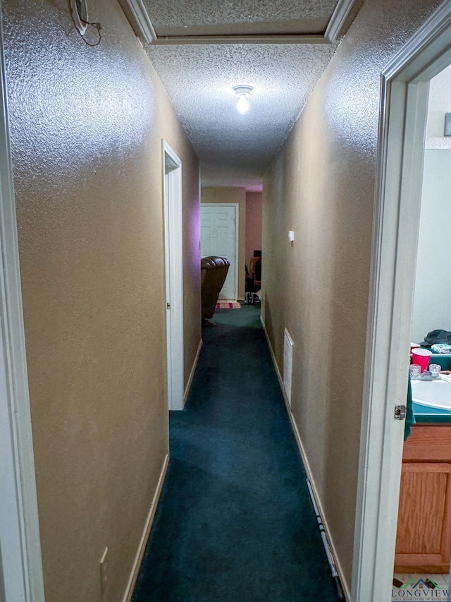 hallway featuring a textured ceiling, dark carpet, attic access, and baseboards