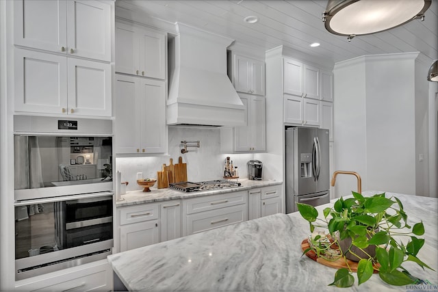 kitchen with light stone countertops, stainless steel appliances, white cabinetry, custom range hood, and backsplash