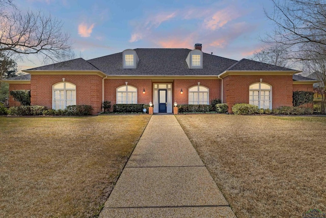 view of front facade with a yard, brick siding, a chimney, and a shingled roof