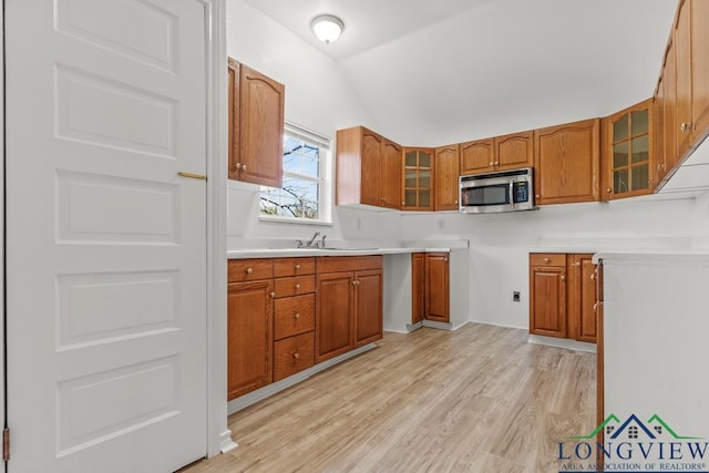 kitchen with stainless steel microwave, glass insert cabinets, vaulted ceiling, brown cabinetry, and a sink