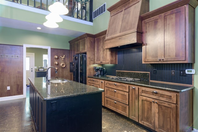 kitchen featuring custom exhaust hood, black appliances, a center island with sink, sink, and dark stone countertops