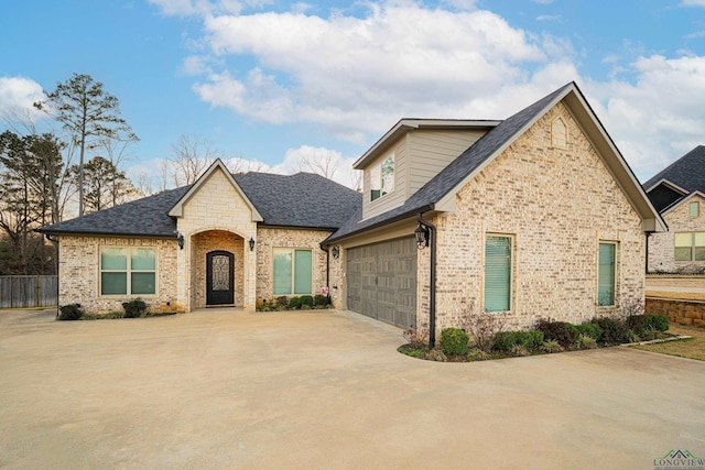 french country style house with an attached garage, brick siding, fence, driveway, and roof with shingles