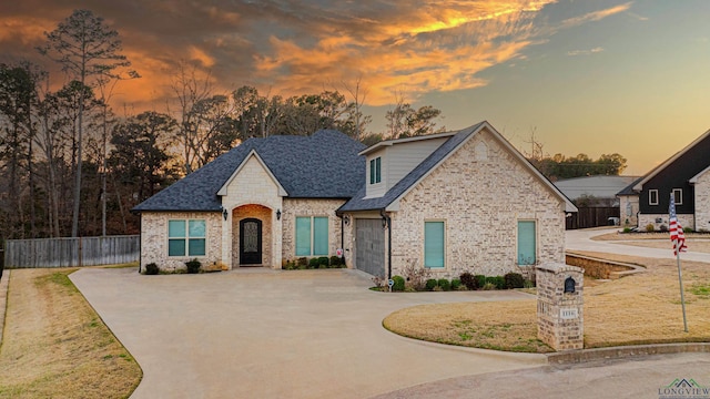 french provincial home featuring concrete driveway, roof with shingles, fence, and brick siding