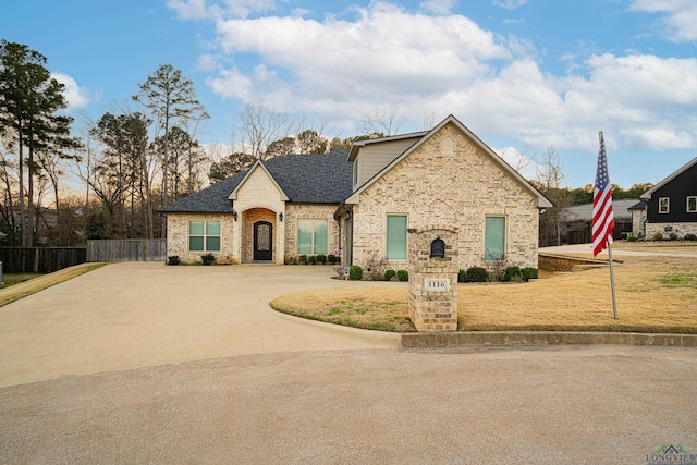 french country inspired facade featuring concrete driveway, roof with shingles, fence, a front lawn, and brick siding