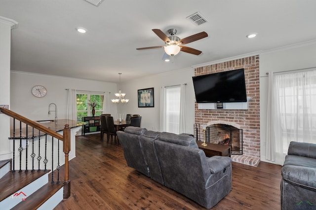 living room with ceiling fan with notable chandelier, ornamental molding, dark wood-type flooring, and a brick fireplace