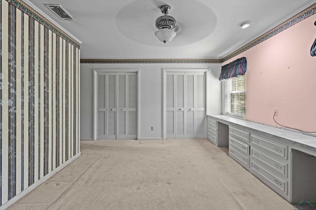 unfurnished bedroom with light carpet, two closets, ceiling fan, built in desk, and ornamental molding