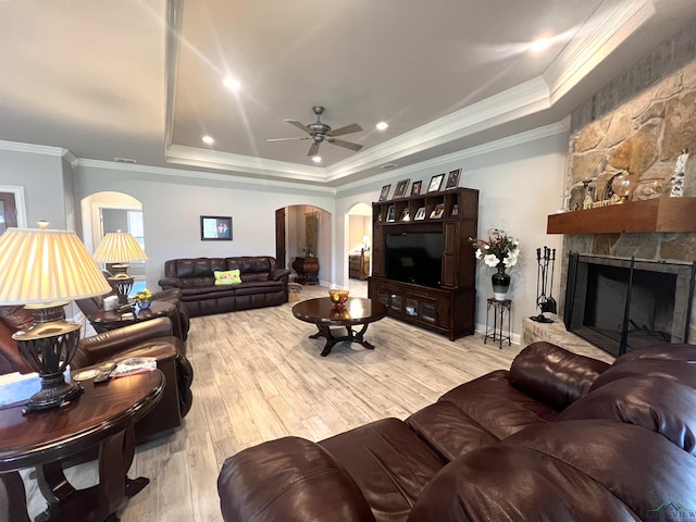 living room featuring ceiling fan, a stone fireplace, crown molding, light hardwood / wood-style floors, and a tray ceiling