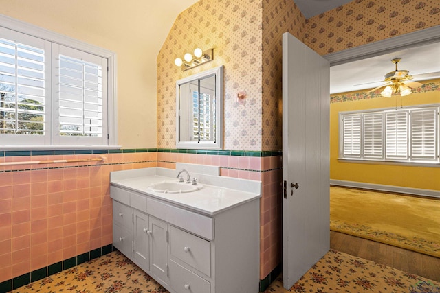 bathroom with wallpapered walls, wainscoting, ceiling fan, vanity, and tile walls