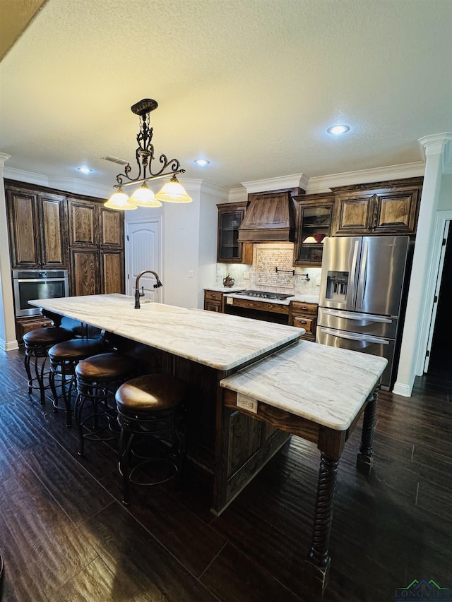 kitchen with appliances with stainless steel finishes, a kitchen island with sink, dark brown cabinets, custom range hood, and pendant lighting