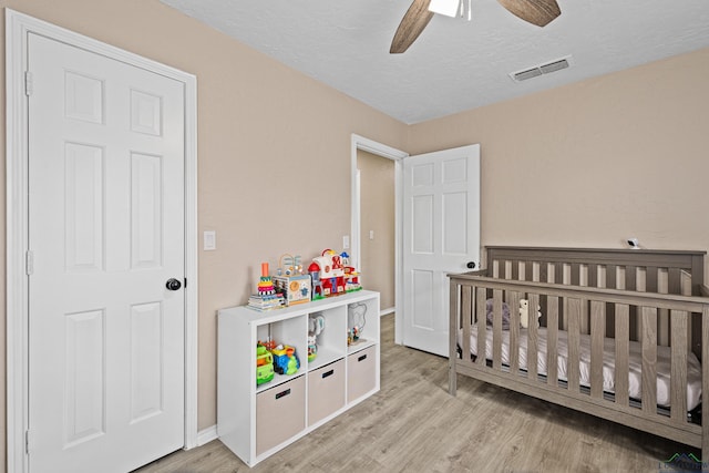 bedroom with ceiling fan, a nursery area, a textured ceiling, and light hardwood / wood-style floors