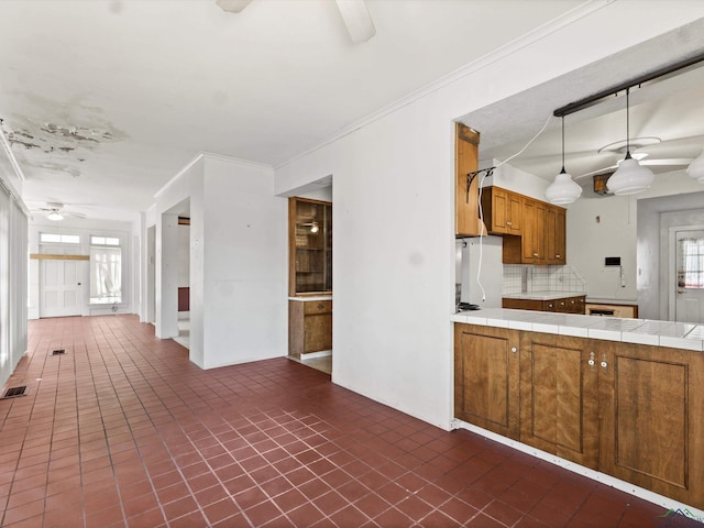 kitchen with tile counters, brown cabinetry, ceiling fan, crown molding, and backsplash