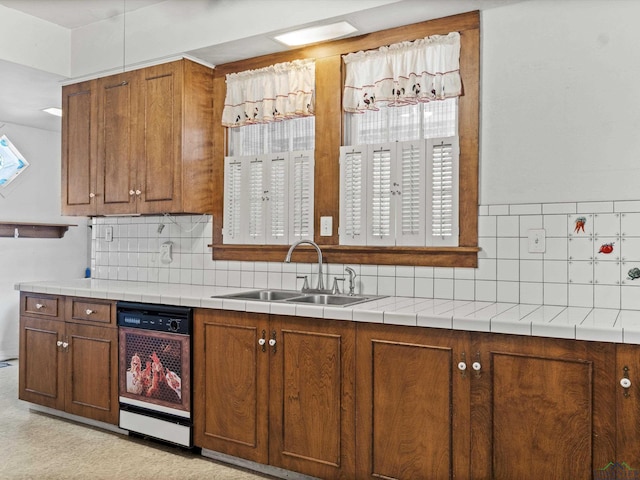 kitchen with brown cabinets, tile countertops, decorative backsplash, a sink, and dishwashing machine