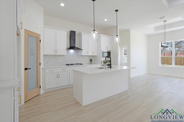 kitchen with wall chimney range hood, sink, white cabinets, a center island with sink, and decorative light fixtures