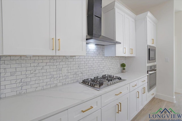 kitchen featuring appliances with stainless steel finishes, white cabinetry, decorative backsplash, light stone countertops, and wall chimney exhaust hood