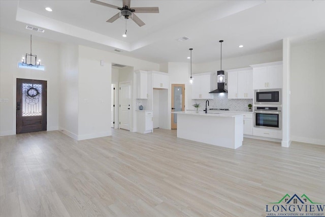 kitchen featuring wall chimney range hood, white cabinetry, an island with sink, built in microwave, and stainless steel oven