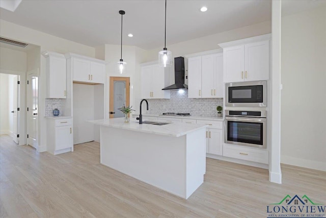 kitchen with sink, appliances with stainless steel finishes, wall chimney range hood, a kitchen island with sink, and white cabinets