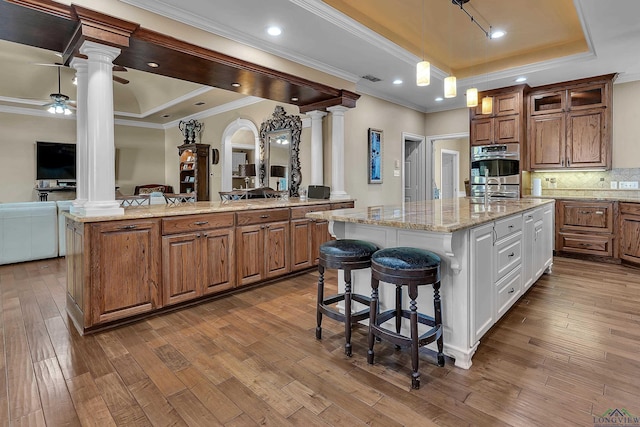 kitchen with ornate columns, ceiling fan, a tray ceiling, a center island with sink, and ornamental molding