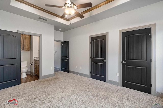 unfurnished bedroom with ensuite bathroom, ornamental molding, a tray ceiling, ceiling fan, and dark colored carpet