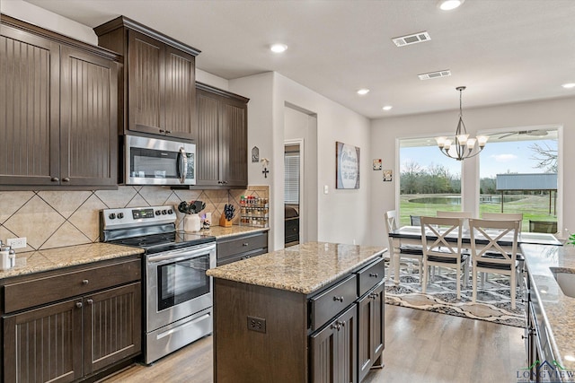 kitchen featuring appliances with stainless steel finishes, a center island, a notable chandelier, light stone counters, and dark brown cabinets