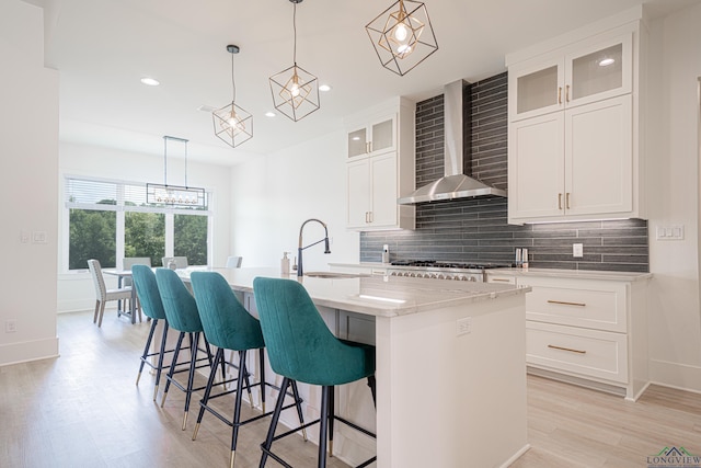 kitchen with stainless steel gas stovetop, wall chimney exhaust hood, an island with sink, decorative light fixtures, and white cabinetry