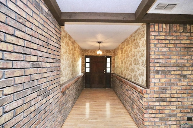 hallway featuring beam ceiling, light hardwood / wood-style flooring, brick wall, and a textured ceiling