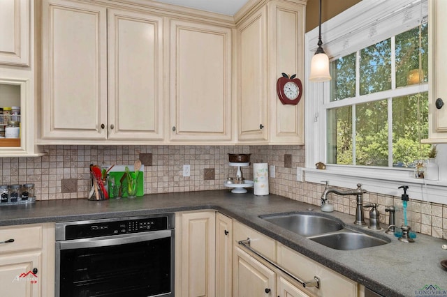 kitchen with stainless steel oven, sink, hanging light fixtures, cream cabinetry, and decorative backsplash