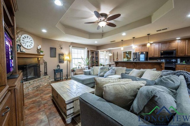 living room with a raised ceiling, ceiling fan, and a brick fireplace