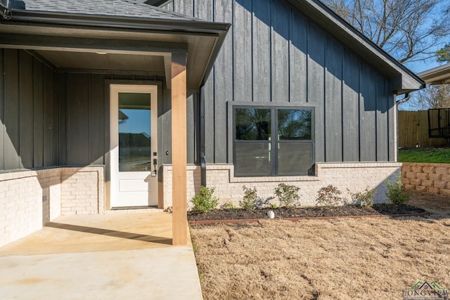 view of exterior entry with brick siding, fence, board and batten siding, and roof with shingles