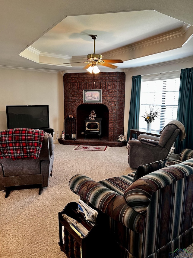 carpeted living room with ceiling fan, a wood stove, ornamental molding, and a tray ceiling