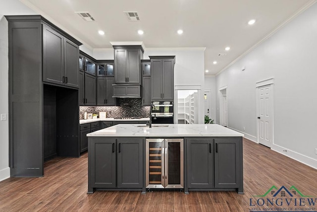 kitchen with dark wood-type flooring, wine cooler, light stone counters, an island with sink, and ornamental molding