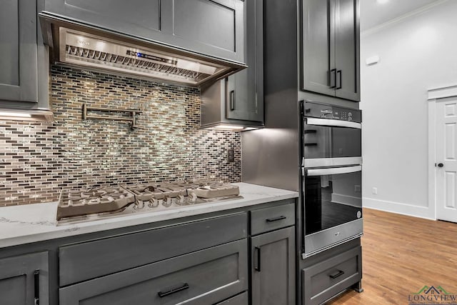 kitchen with stainless steel gas stovetop, exhaust hood, multiple ovens, decorative backsplash, and light stone countertops