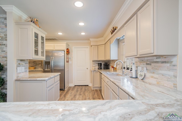 kitchen featuring a sink, light stone counters, stainless steel fridge with ice dispenser, crown molding, and glass insert cabinets