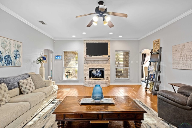 living room featuring light wood finished floors, visible vents, arched walkways, crown molding, and a brick fireplace