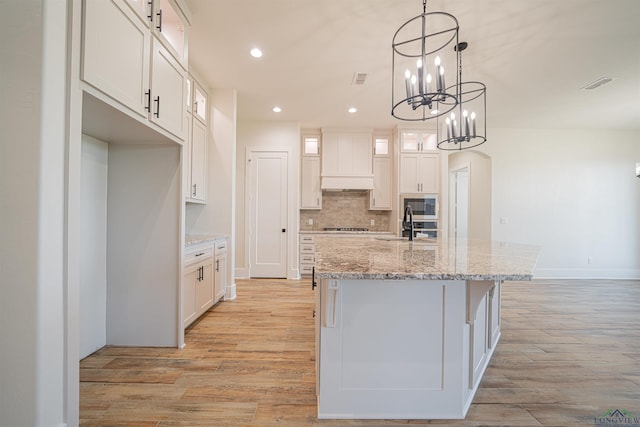 kitchen with custom exhaust hood, a center island with sink, light hardwood / wood-style flooring, light stone counters, and white cabinetry