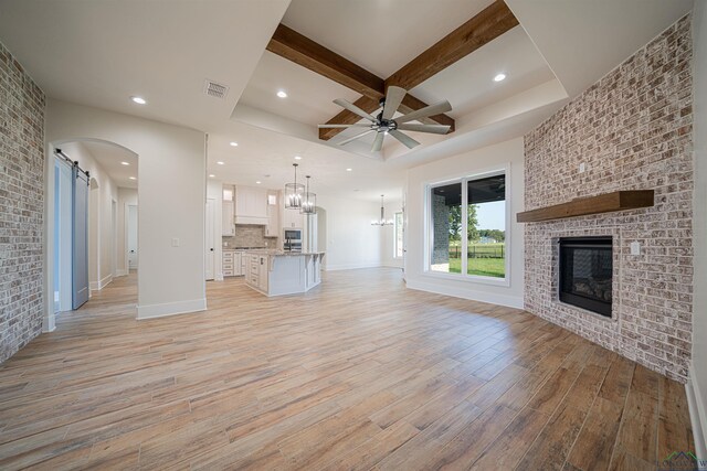 unfurnished living room featuring beam ceiling, a barn door, a fireplace, ceiling fan with notable chandelier, and light wood-type flooring